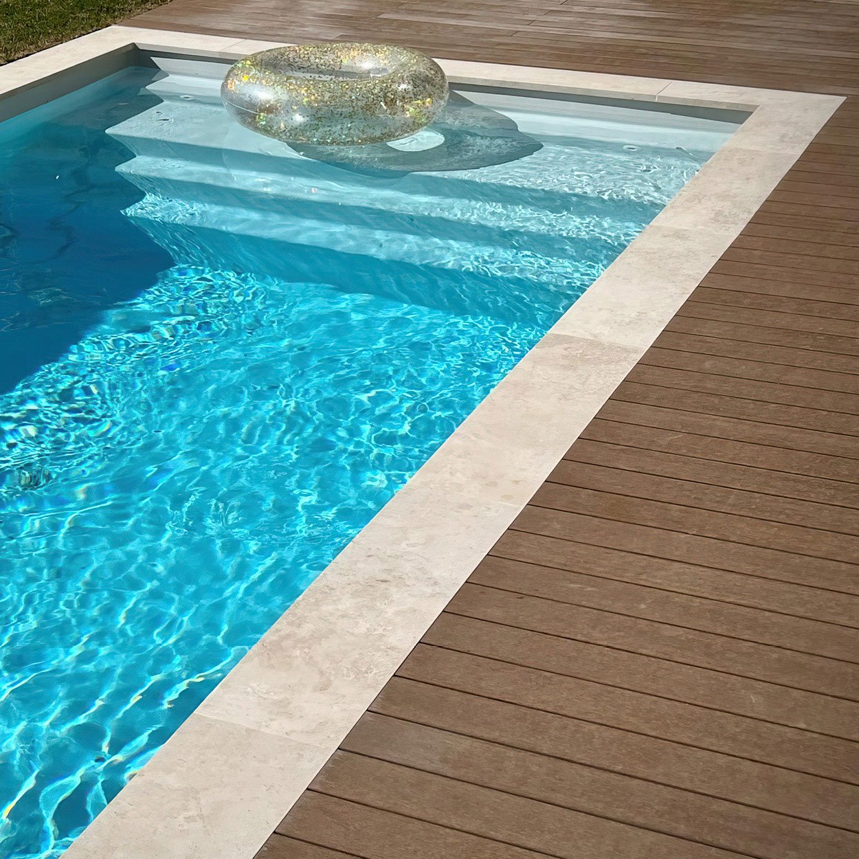Piscine coque aux contours arrondis entourée d'une terrasse en bois, eau bleu clair avec reflets solaires et un flotteur transparent, située dans un espace détente avec margelles en pierre et végétation en arrière-plan.