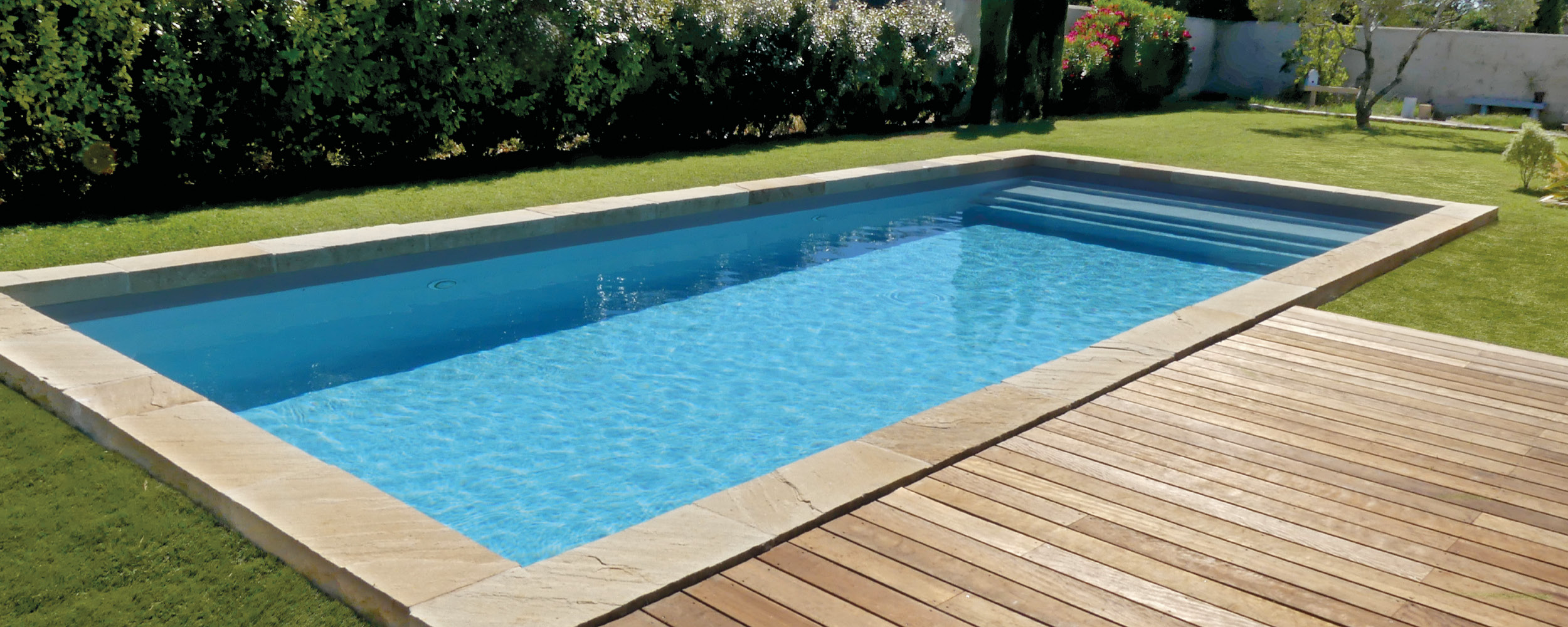 Piscine coque rectangulaire moderne dans un jardin avec pelouse et terrasse en bois, entourée de haies, sous un ciel ensoleillé.