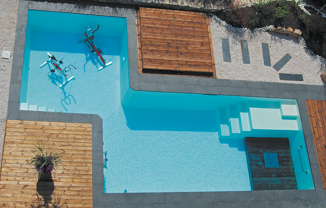 Piscine coque moderne avec forme rectangulaire et coins arrondis, équipée de zones de profondeur variées et de marches intégrées pour un accès facile. Aménagement extérieur incluant une terrasse en bois, margelle en béton gris, éléments de décoration tels qu'une plante en pot et équipements de fitness aquatique, entourée de gravier et de pierres décoratives, dans un environnement lumineux et naturel.
