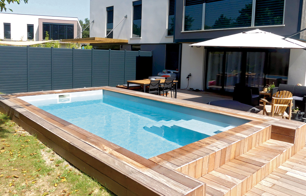 Piscine coque moderne rectangulaire avec marches intégrées, entourée d'un bord en bois chaleureux et sécurisé par un treillis gris foncé, aménagée avec mobilier de jardin confortable et parasol, devant une maison au design contemporain avec grandes fenêtres, le tout dans un jardin minimalistement paysagé.