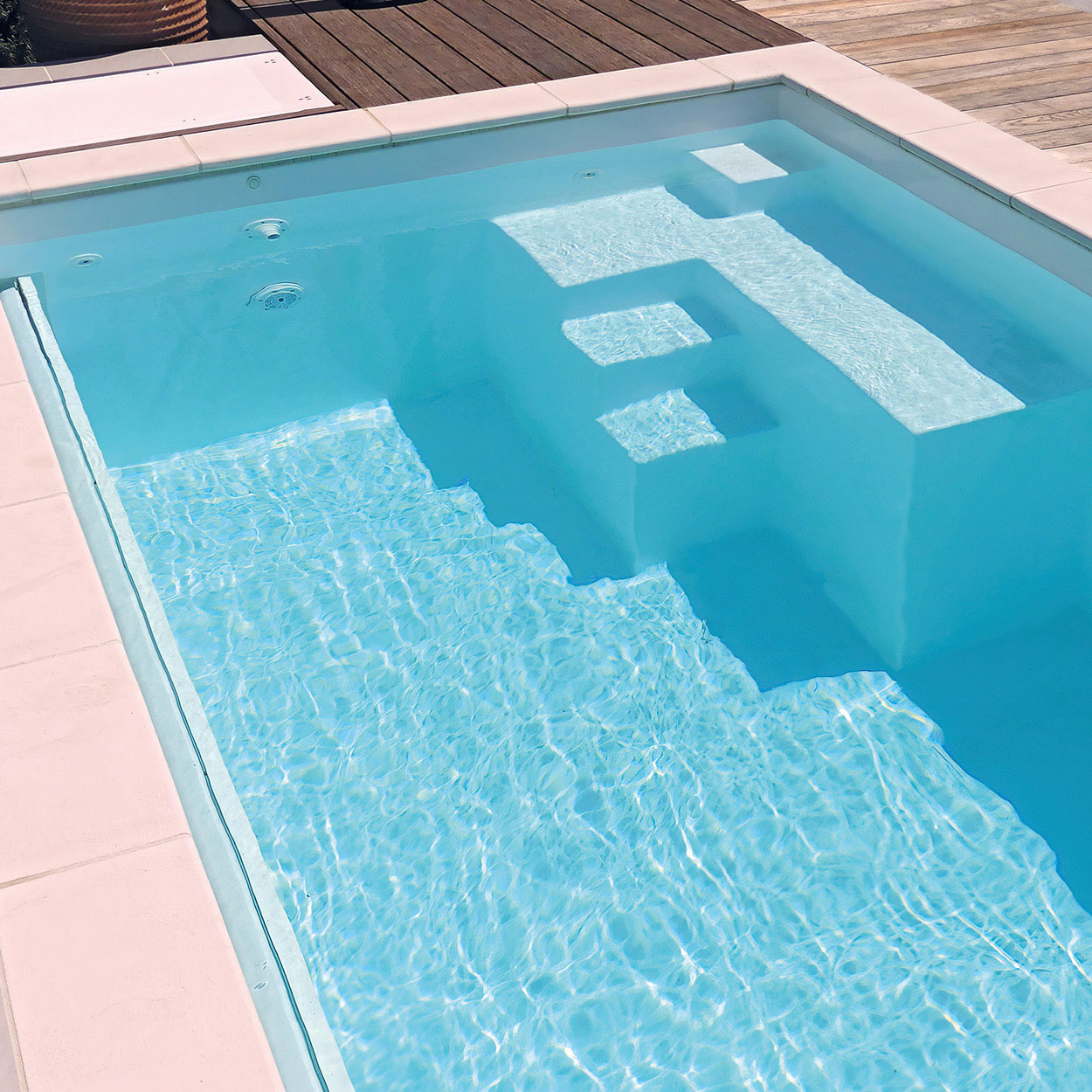 Piscine coque bleu clair avec design géométrique et marches intégrées, entourée de revêtement en bois, offrant une ambiance rafraîchissante et zen avec eau limpide, idéale pour détente et entretien facile.