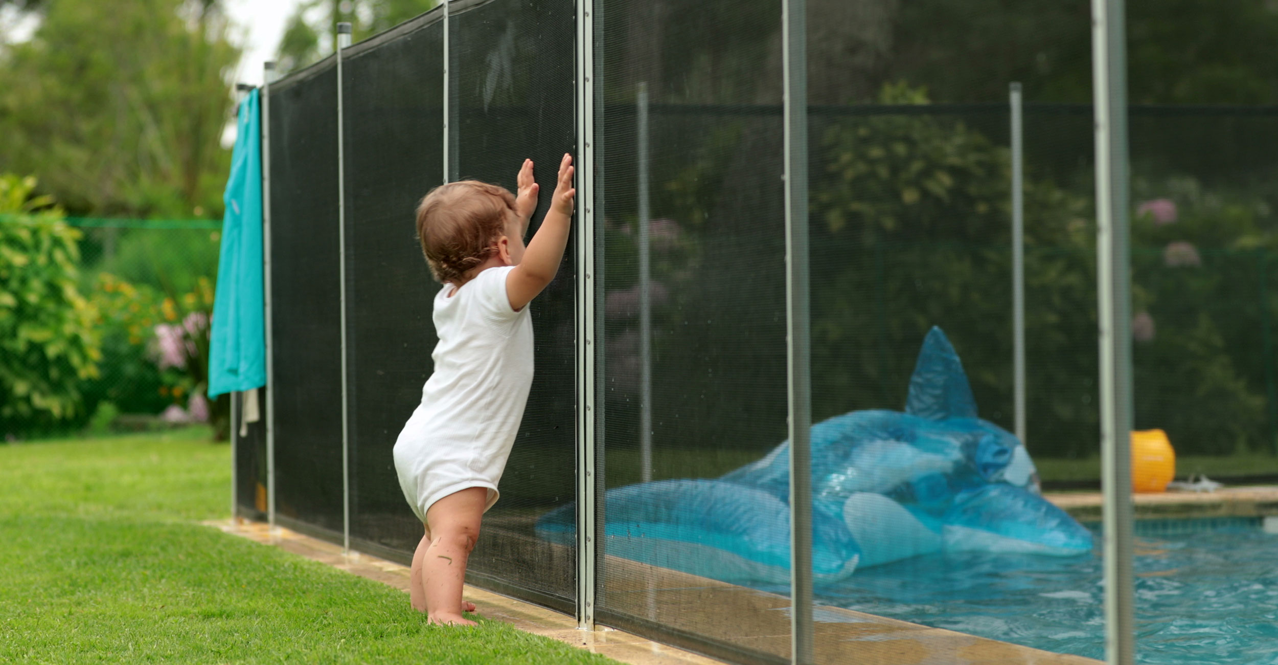 Enfant curieux observant une piscine coque sécurisée avec un jouet requin flottant, entourée de verdure dans un jardin familial