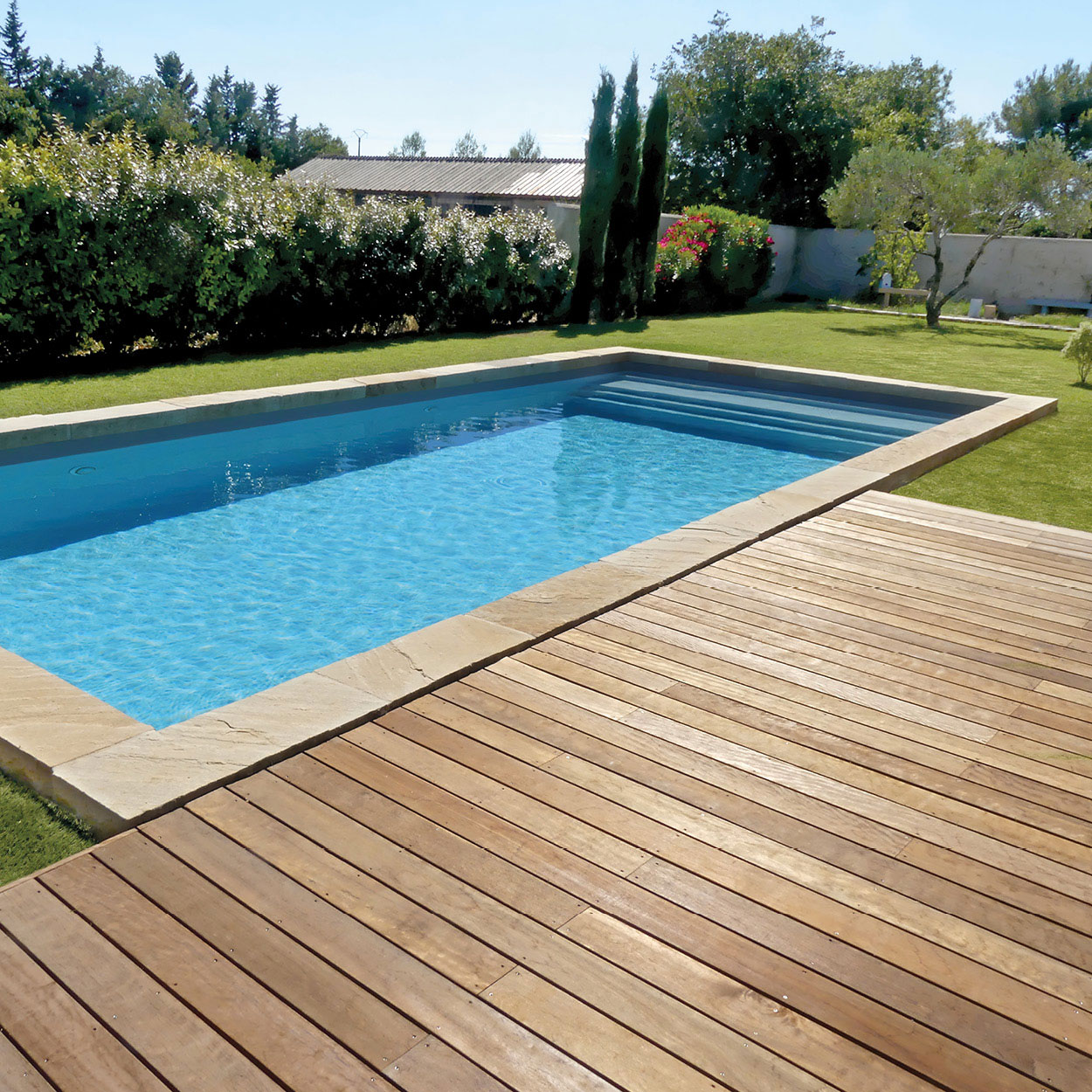 Piscine coque rectangulaire aux bords arrondis dans un jardin avec terrasse en bois, pelouse verte, plantes fleuries et arbres, sous une lumière naturelle