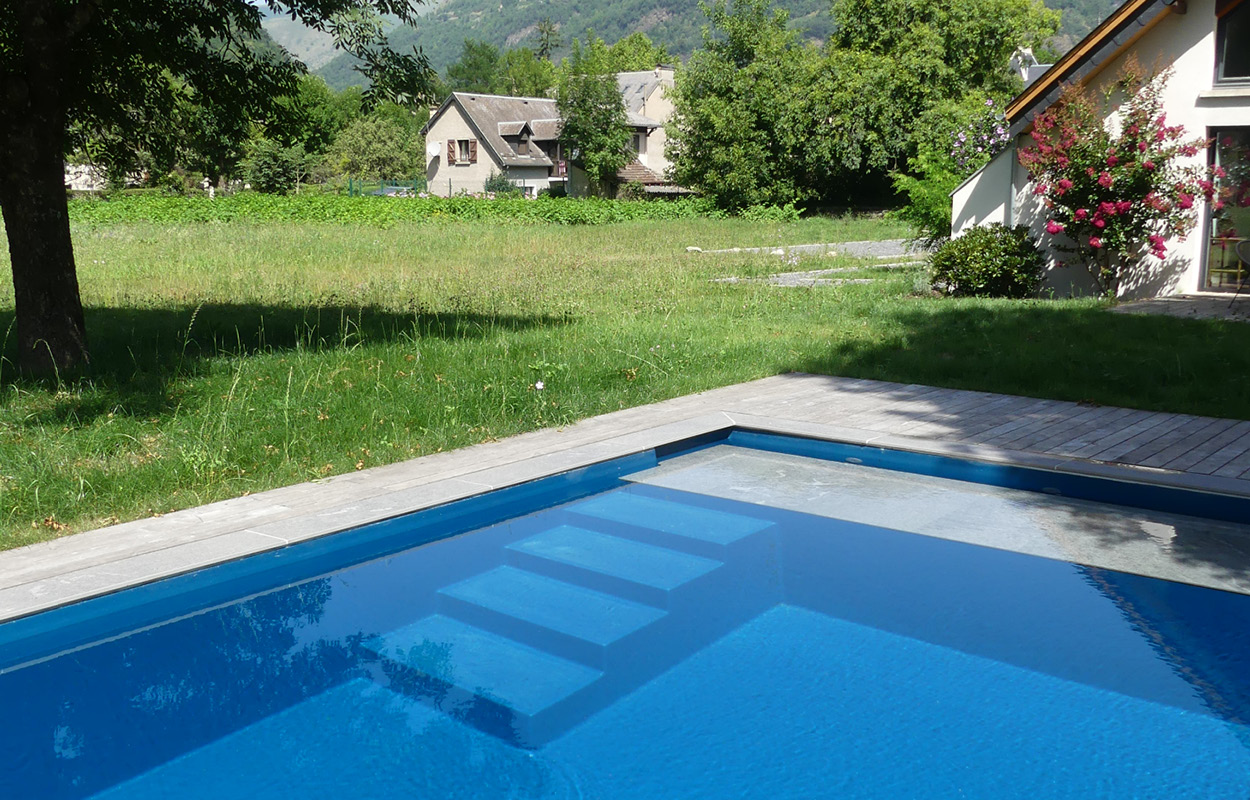 Piscine coque moderne et fonctionnelle avec marches intégrées, entourée de verdure et d'une pelouse bien entretenue, dotée d'un contour en pierre ou carrelage élégant, située devant des maisons traditionnelles et embellie par des fleurs, reflétant une lumière naturelle dynamique.