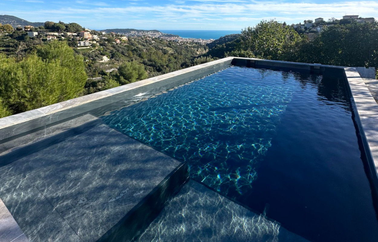 Piscine à débordement avec design moderne et épuré, s'intégrant harmonieusement dans un environnement naturel avec vue sur la mer. Lignes élégantes, surface claire et réfléchissante, entourée de verdure luxuriante, idéale pour détente et intégration paysagère. Style de luxe pour piscines coque.