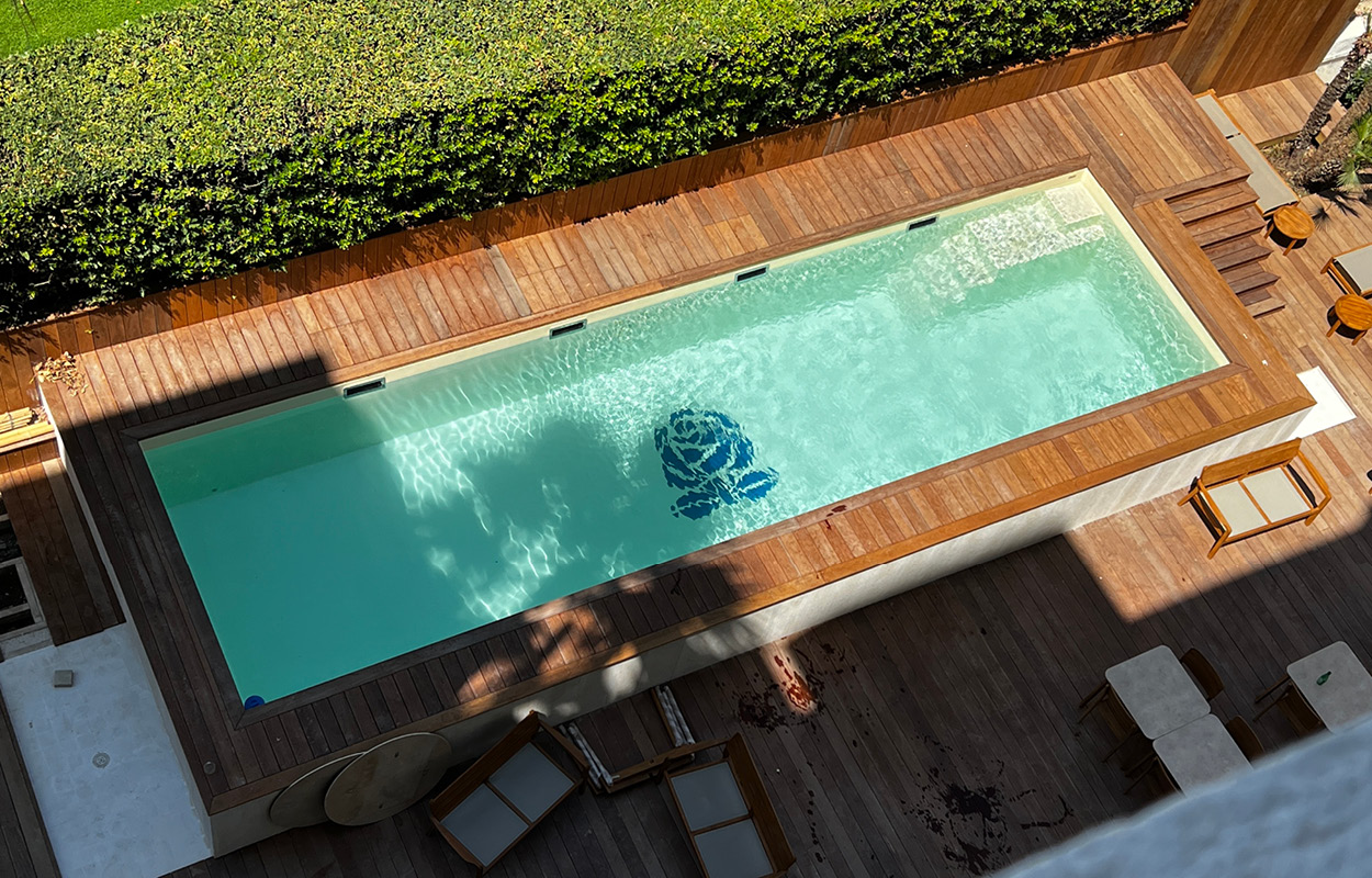Piscine coque rectangulaire en polyester entourée d'un deck en bois avec ambiance chaleureuse, eau turquoise claire, haie verdoyante, mobilier de détente, motif de rose au fond et éclairage naturel.