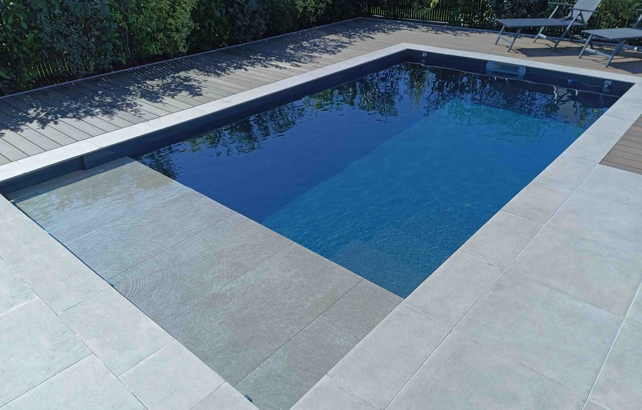 Piscine coque moderne avec surface en gelcoat brillant entourée de dalles grises antidérapantes, eau turquoise claire, terrasse en bois, mobilier de jardin pour la détente et verdure en arrière-plan.