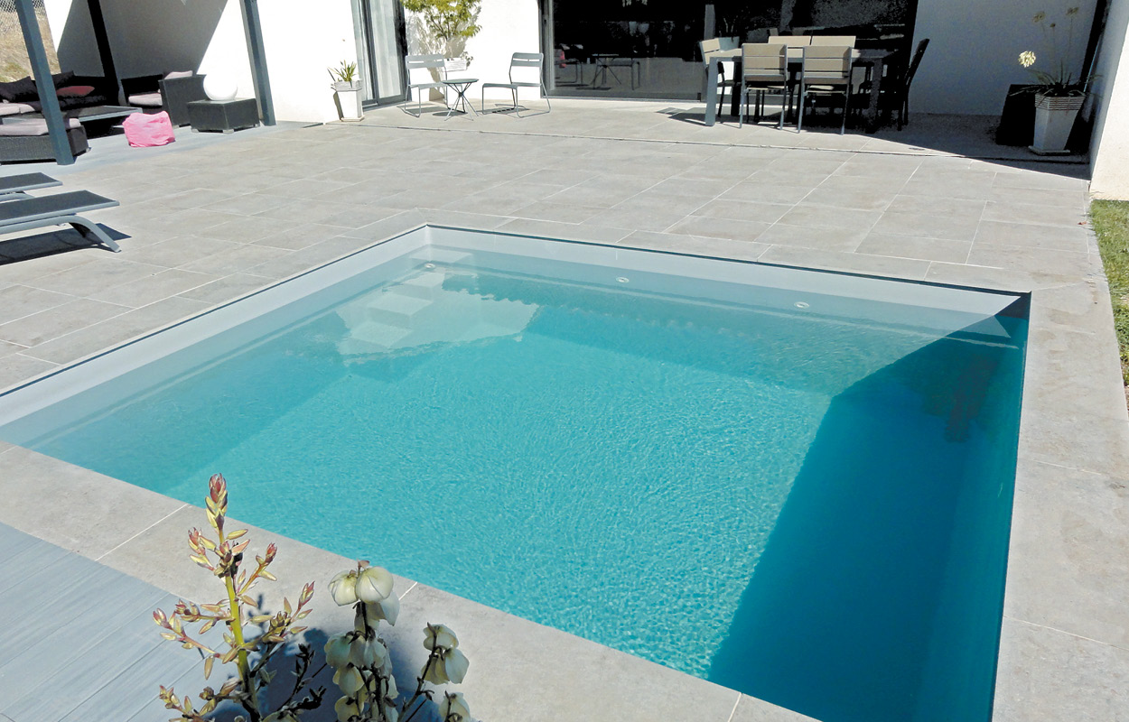 Piscine coque rectangulaire moderne avec eau turquoise, entourée de carrelage clair et plantes décoratives, espace détente avec transats et mobilier d'extérieur pour un cadre accueillant et bien aménagé.
