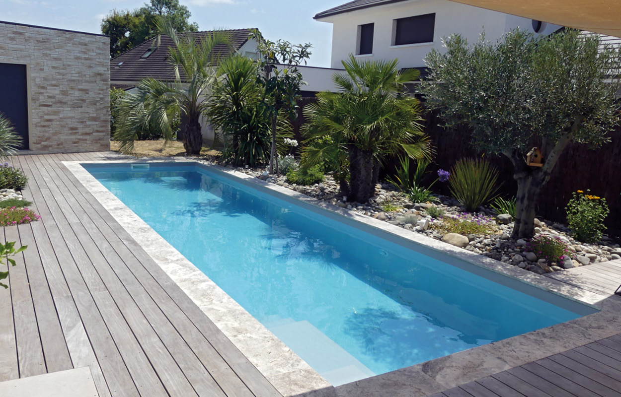 Piscine coque rectangulaire avec margelles en pierre naturelle et terrasse en bois, entourée de palmiers et de plantes exotiques dans un jardin bien aménagé.