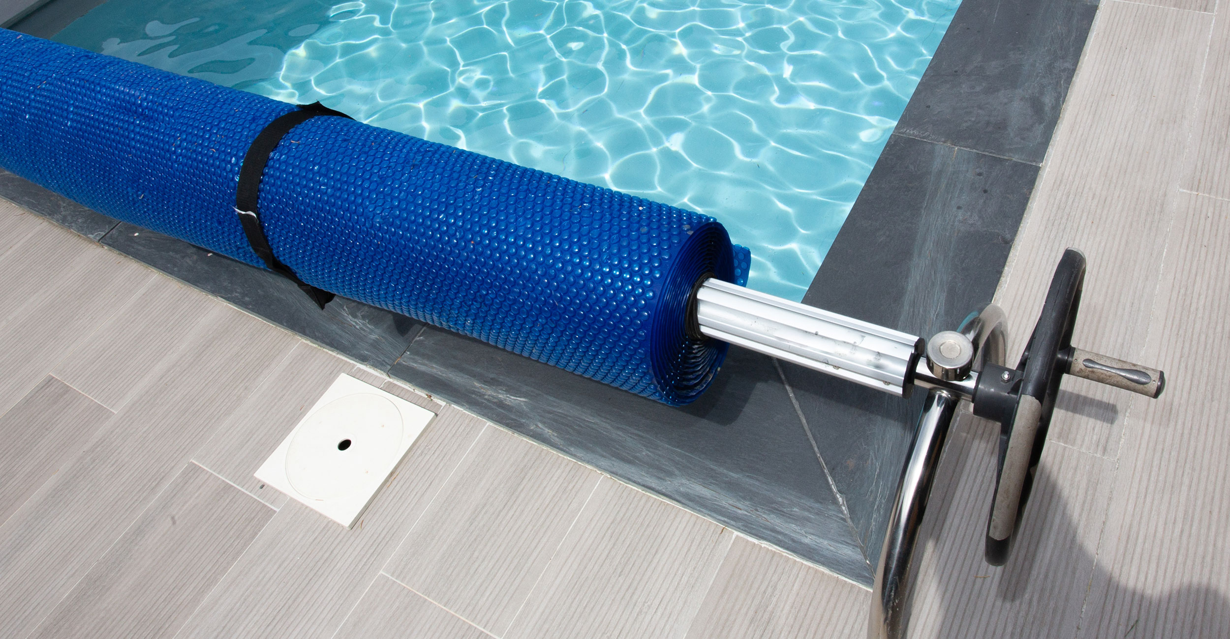 Piscine coque avec eau turquoise et enrouleur de couverture à manivelle, entourée de dalles modernes en plein soleil