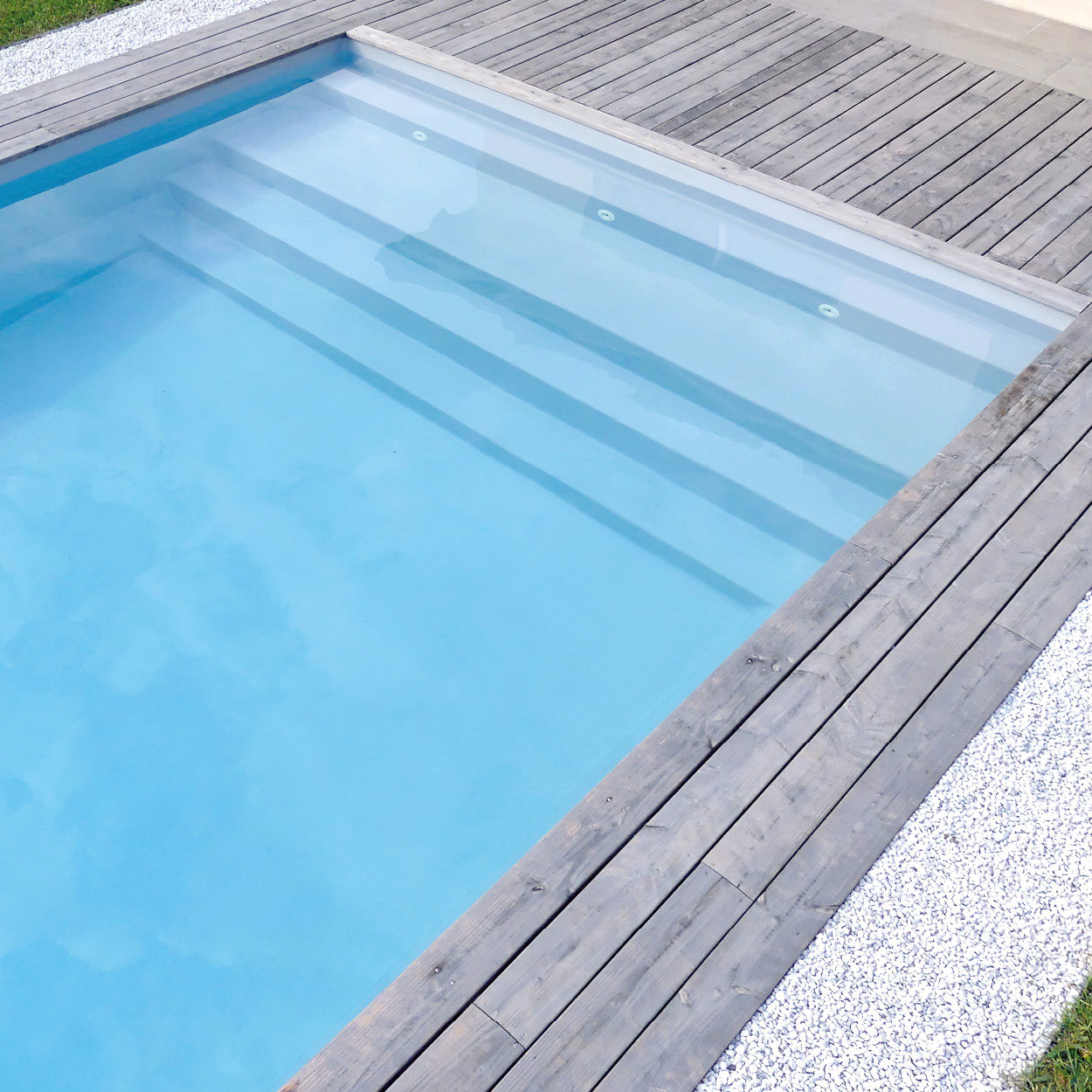 Piscine coque moderne avec finition lisse et parois en plastique renforcé, entourée de bordures en bois naturel et graviers blancs, eau bleu clair translucide, éclairage intégré pour ambiance nocturne, design épuré et espace extérieur élégant, idéal pour amateurs de loisirs aquatiques et design extérieur.