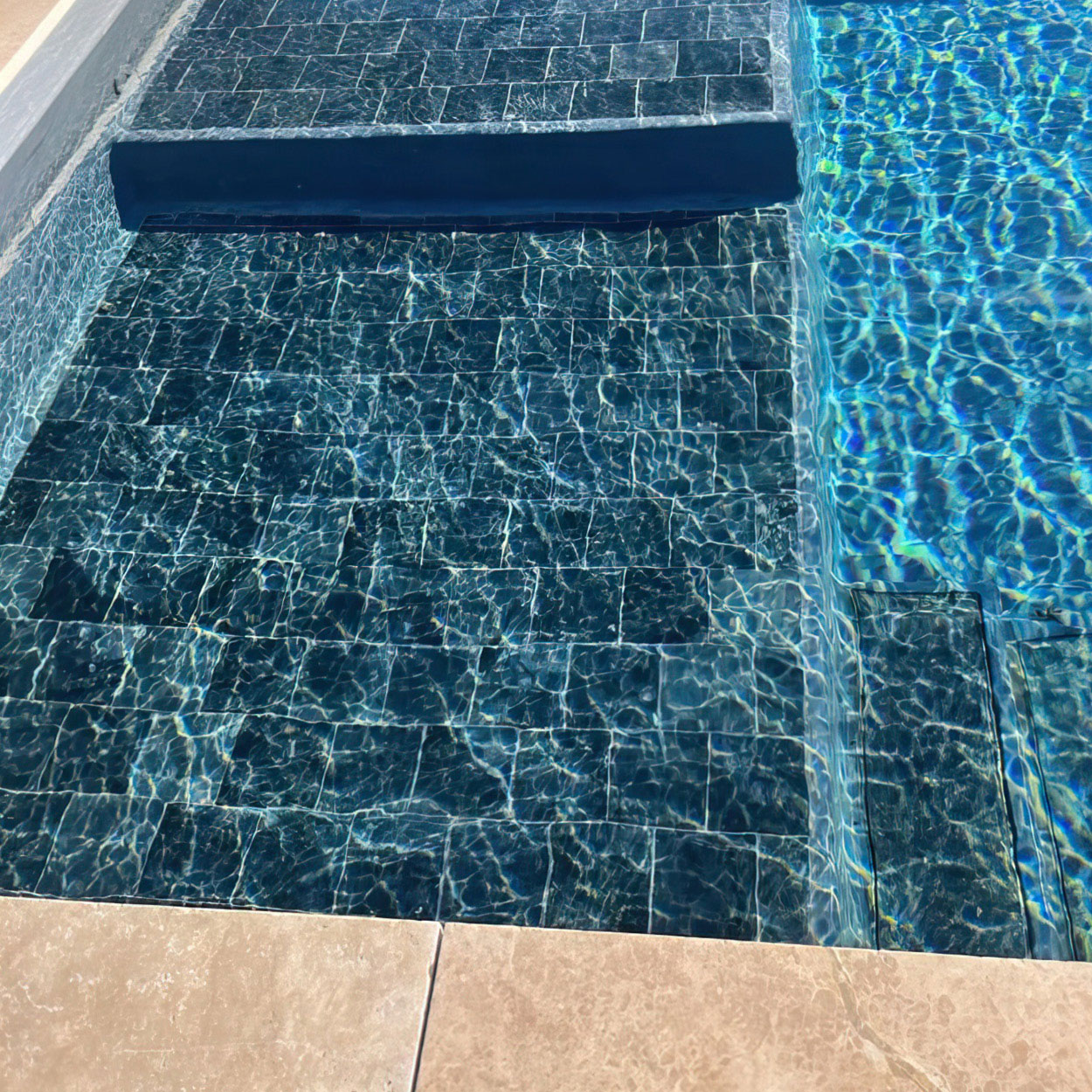 Piscine coque moderne avec fond en carreaux sombres, eau claire et reflets lumineux, escalier intégré pour accès sécurisé, bordure beige en pierre, idéale pour détente et loisir.