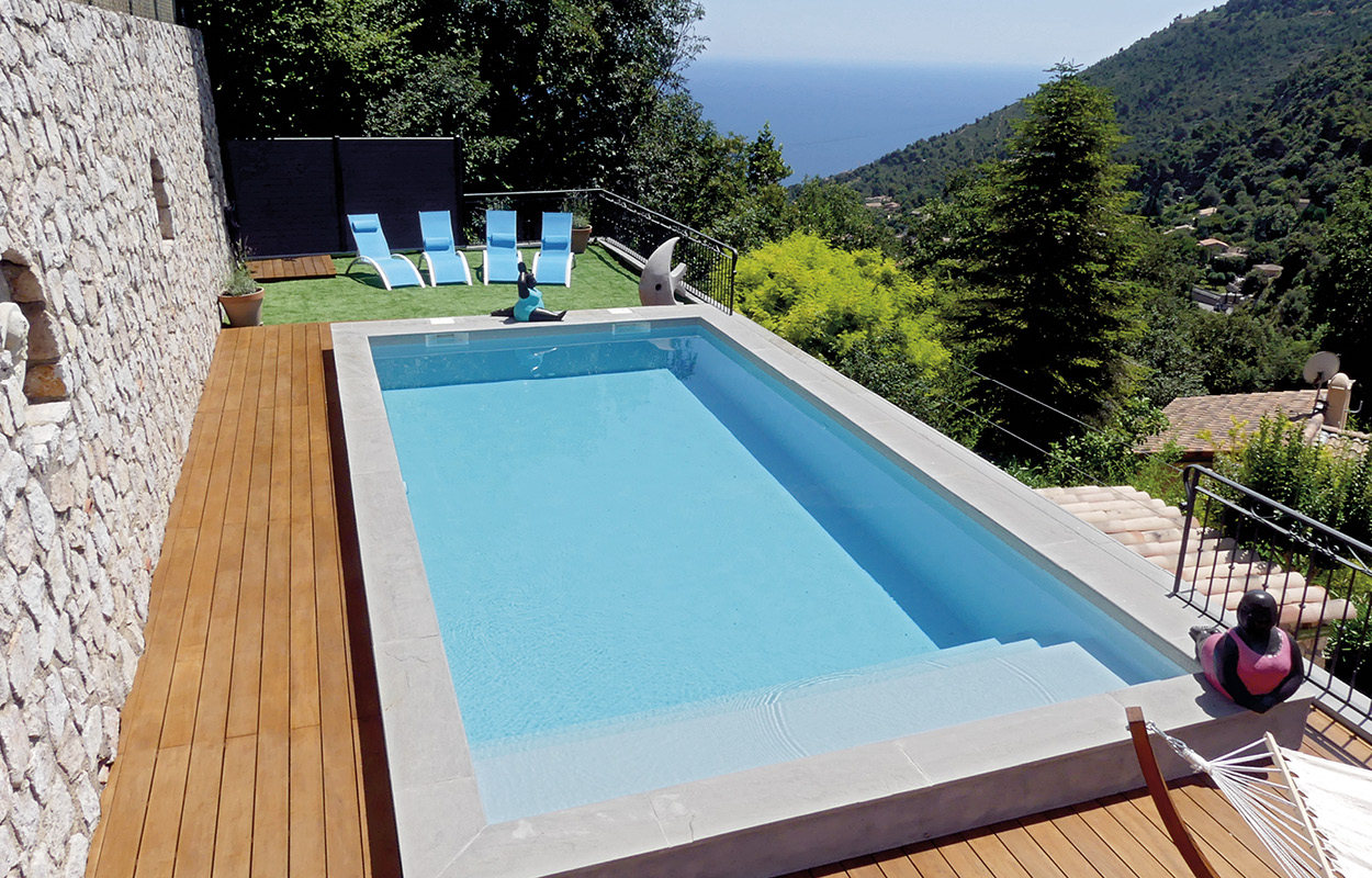 Espace extérieur avec piscine coque rectangulaire entourée de terrasse en bois, trois chaises longues bleues sur pelouse avec vue panoramique montagne et mer, éclairage naturel ensoleillé, aménagement élégant et serein parfait pour détente.