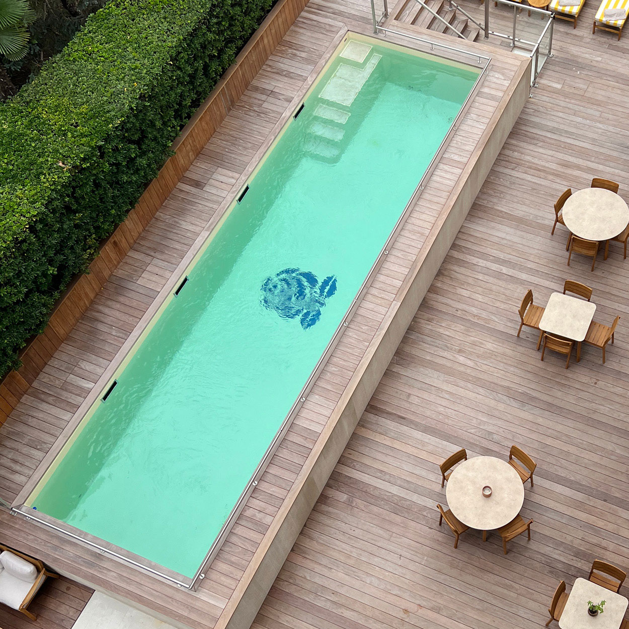 Piscine coque rectangulaire avec eau vert clair, design moderne avec decking en bois, escalier en béton, motif de mosaïque, mobilier d'extérieur en bois, végétation luxuriante pour un espace de détente luxueux et convivial.