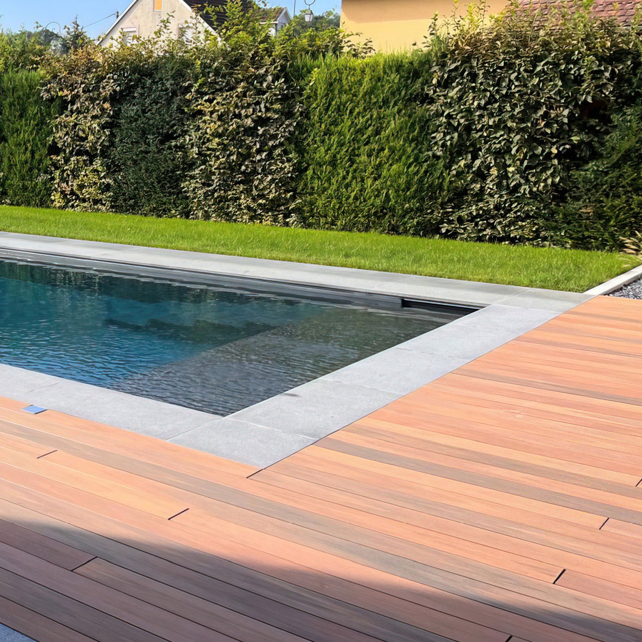 Installation élégante de piscine coque avec aménagement paysager soigné, présentant une eau claire et réfléchissante, entourée d'une margelle en pierre grise et d'un platelage en bois chaleureux, accompagnée d'une haie d'arbustes taillés pour l'intimité et d'un espace vert bien entretenu, créant une atmosphère naturelle et accueillante idéale pour la détente en plein air.