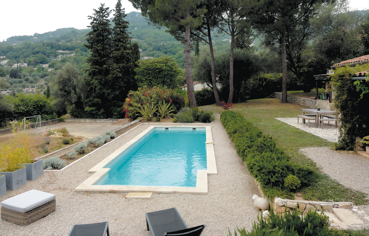 Piscine coque rectangulaire au design moderne, intégrée dans un jardin soigné avec gravier, plantes et arbres, entourée de mobilier de jardin confortable, offrant une vue sur des collines verdoyantes.