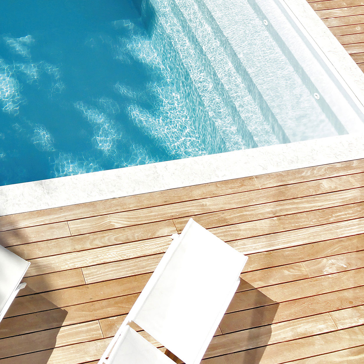 Piscine coque moderne et élégante à l'eau bleu éclatant, entourée d'un revêtement en pierre clair et d'un deck en bois haut de gamme, avec chaises longues blanches pour la détente, sous un plein ensoleillement, reflétant propreté et sérénité.
