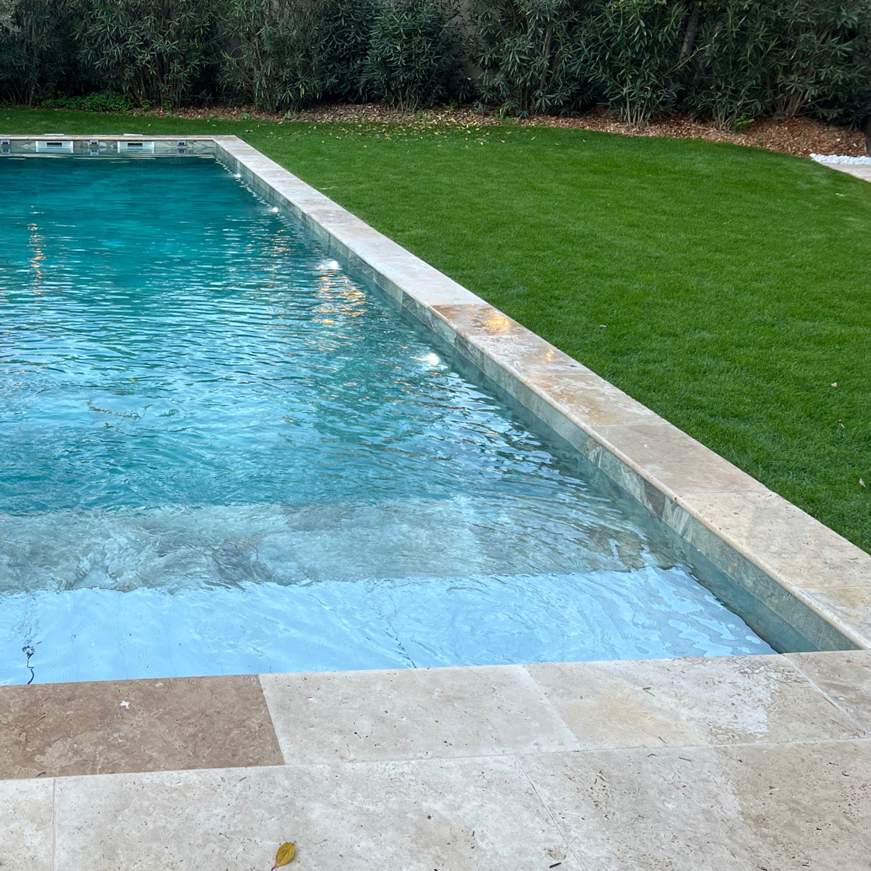 Piscine coque bleu clair avec margelles en pierre dans un cadre paysagé, entourée de pelouse verte et haies pour l'intimité, éclairage intégré pour les soirées, idéale pour un espace de détente extérieur