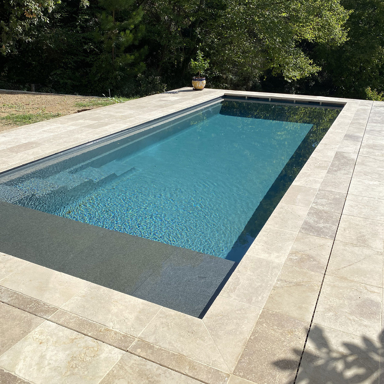 Piscine coque moderne à débordement entourée d'une terrasse avec dalles de pierre, et margelle sombre, dans un cadre naturel avec arbres. Escalier intégré et pot décoratif visibles.