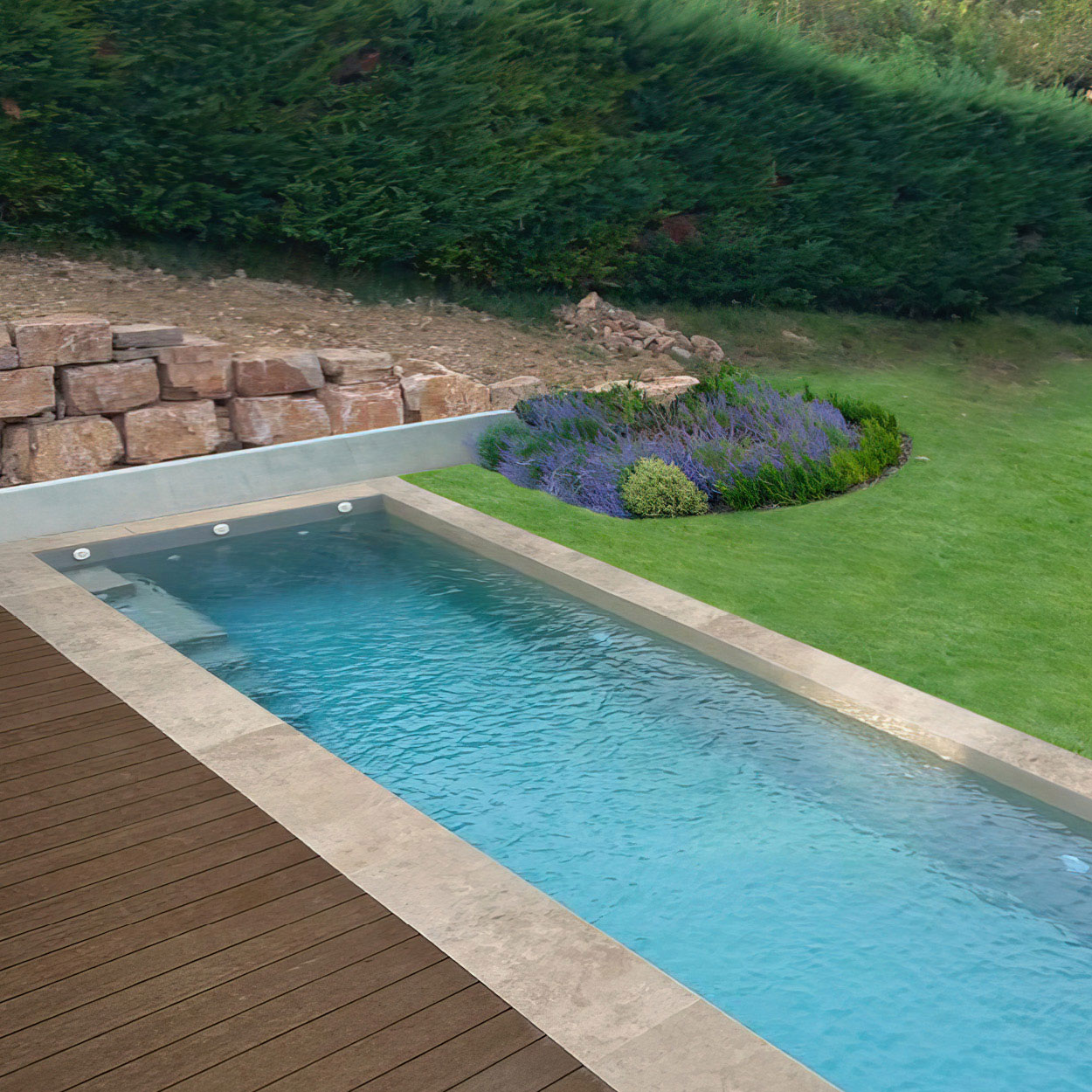 Piscine coque rectangulaire moderne avec parois claires entourée de lavande et d'une terrasse en bois dans un jardin aménagé avec pelouse et mur en pierre, créant un espace extérieur élégant et relaxant