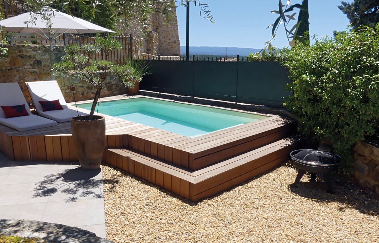 Piscine coque rectangulaire moderne entourée d'un deck en bois avec mobilier confortable et végétation décorative, aménagement extérieur harmonieux et sécurisé pour détente en journée ensoleillée.