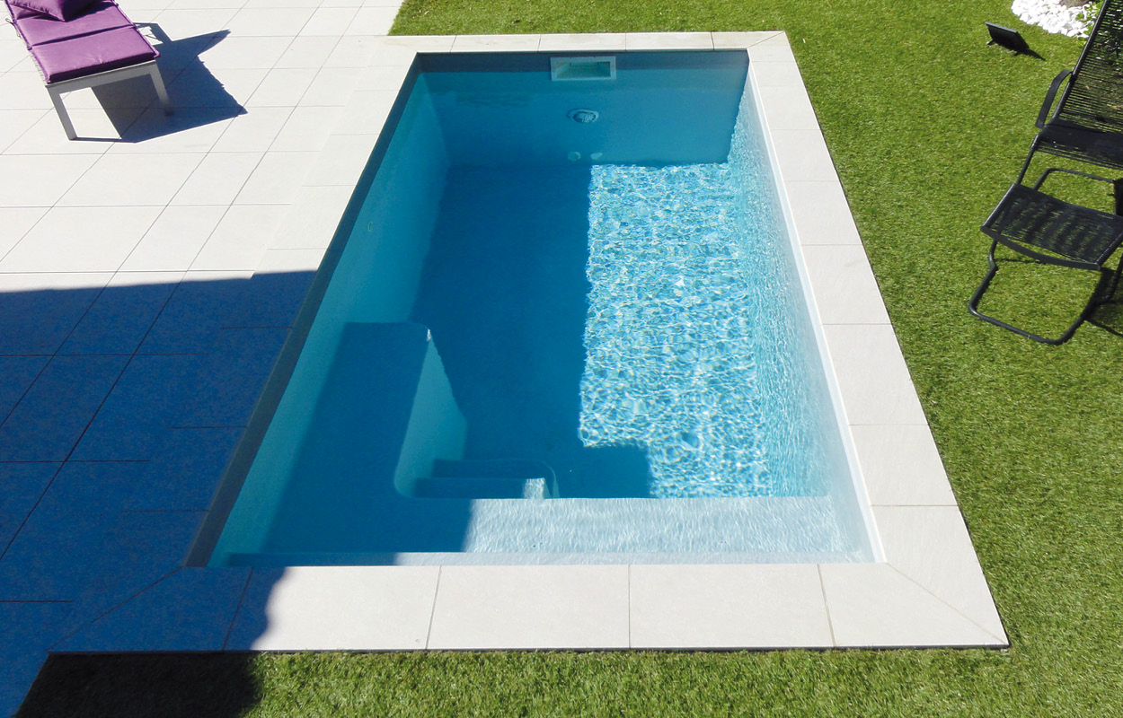 Piscine coque moderne avec angles arrondis, entourée de carrelage clair et gazon artificiel, transat violet à gauche, idéale pour détente extérieure