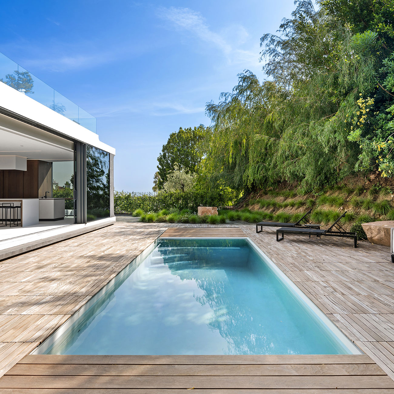Espace extérieur contemporain avec piscine coque rectangulaire, terrasse en bois, design architectural moderne avec baies vitrées, végétation luxuriante, mobilier extérieur élégant, ciel dégagé, ambiance luxe et confort.