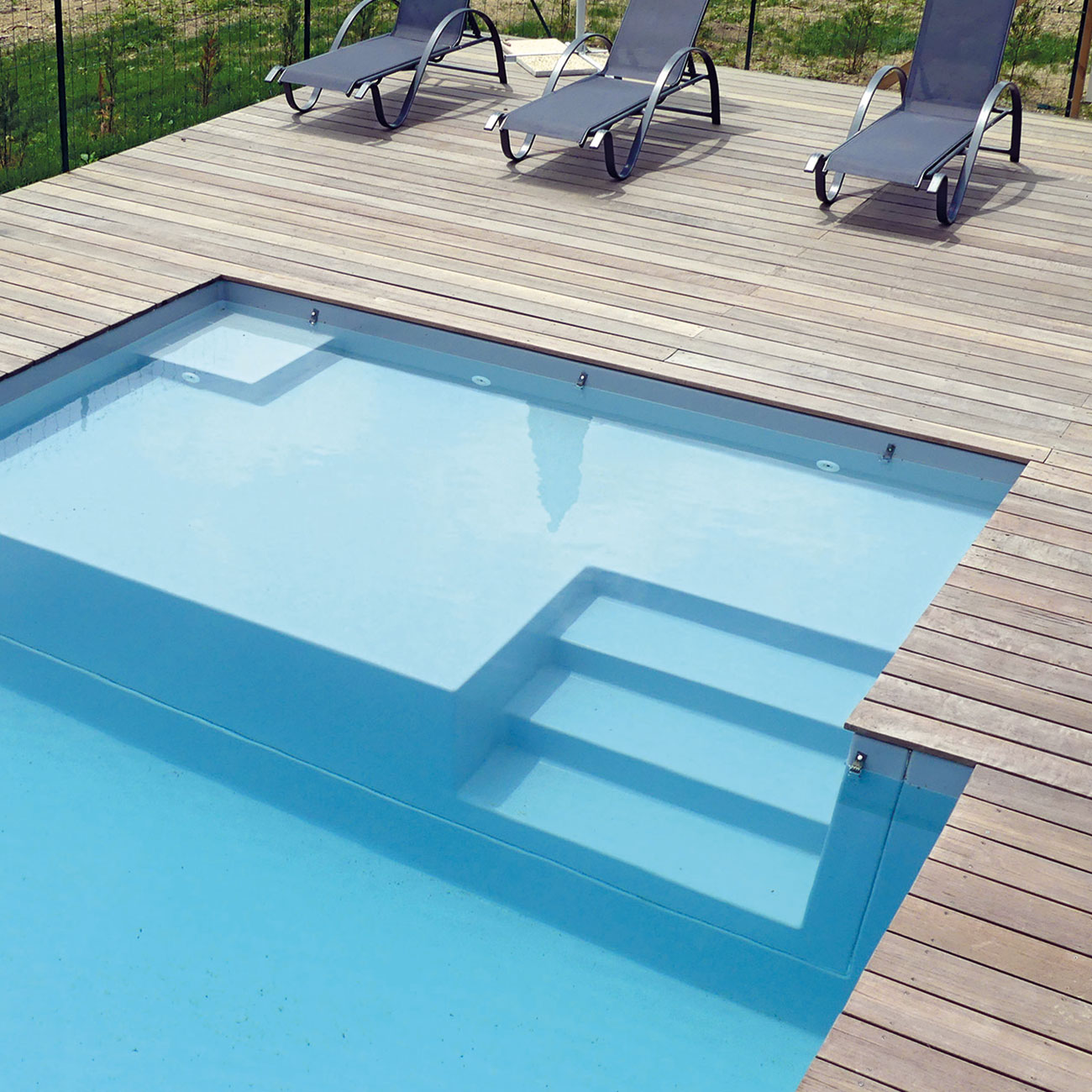 Piscine coque moderne avec escaliers intégrés, eau bleu clair reflétant le soleil, terrasse en bois chaleureux et chaises longues design, entourée d'espaces verts pour une ambiance détente et relaxante.