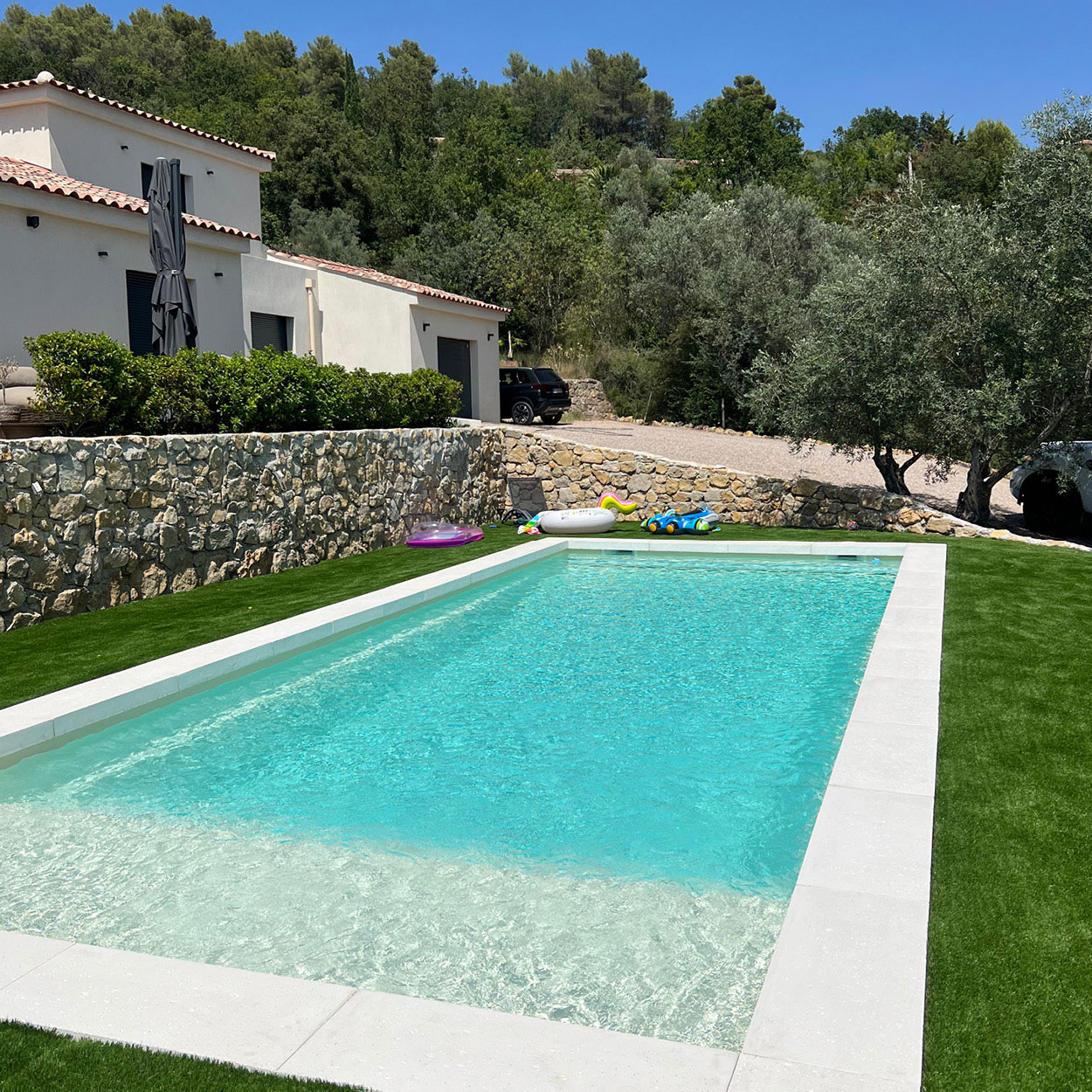 Piscine coque spacieuse dans un cadre naturel avec maison moderne, bordures en béton clair, eau turquoise claire, gazon synthétique et mur en pierre, offrant détente et confort près de collines verdoyantes.