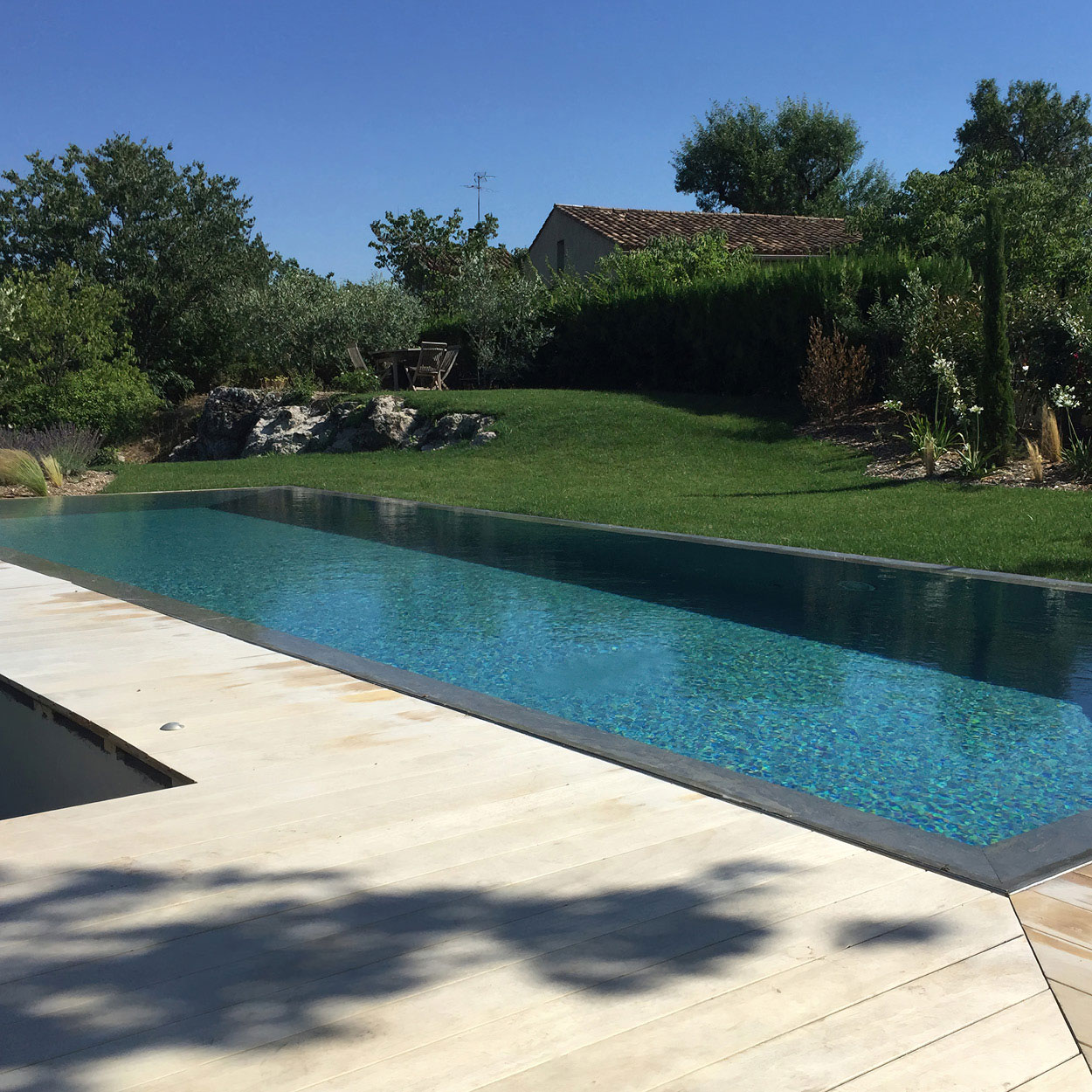 Piscine coque rectangulaire turquoise avec contour béton moderne et plage en bois, entourée de verdure et aménagement paysager soigné avec herbe, rochers, arbres, et mobilier de jardin sous ciel ensoleillé.