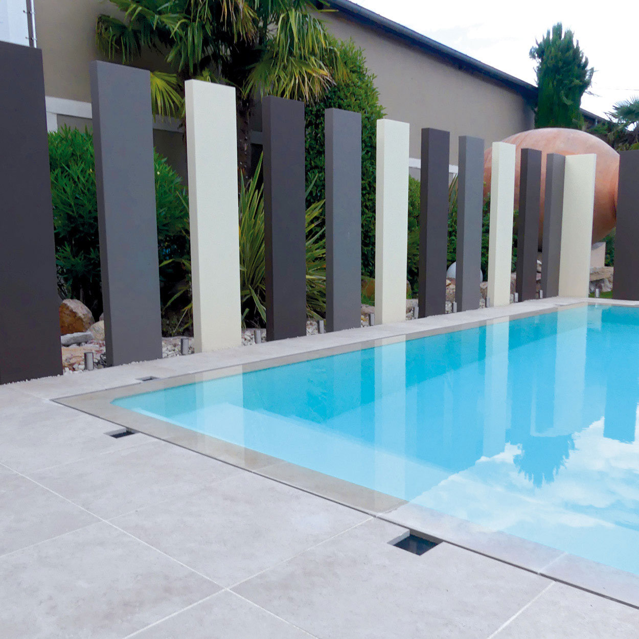 Piscine coque moderne rectangulaire entourée d'un aménagement paysager soigné avec palmiers, fleurs ornementales et revêtement de sol en carrelage clair, dans un espace extérieur harmonieux et accueillant.