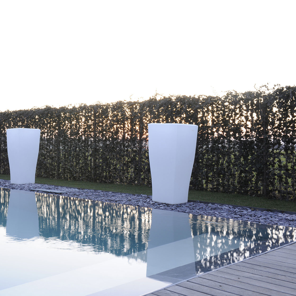 Espace extérieur moderne et sophistiqué avec piscine coque minimaliste entourée de dalles en pierre naturelle, grands pots blancs décoratifs, mur végétal pour l'intimité, et éclairage doux pour ambiance luxueuse et détente