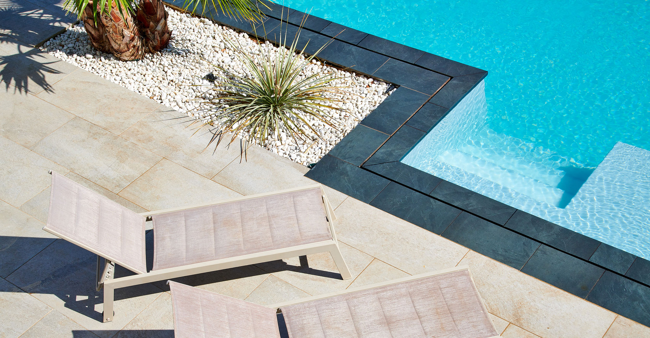 Piscine coque moderne entourée d'une terrasse accueillante avec dalles sombres, chaises longues minimalistes, plantes tropicales et ambiance luxueuse, idéale pour la détente.
