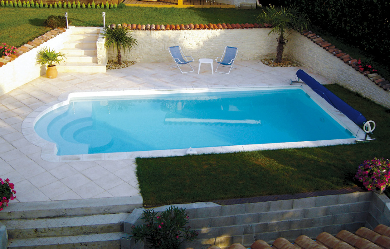 Piscine coque ovale avec eau bleu clair, entourée de dalles en pierre et pelouse, mobilier de jardin avec chaises longues, plantes tropicales et fleurs colorées, espace détente extérieur élégant et apaisant.