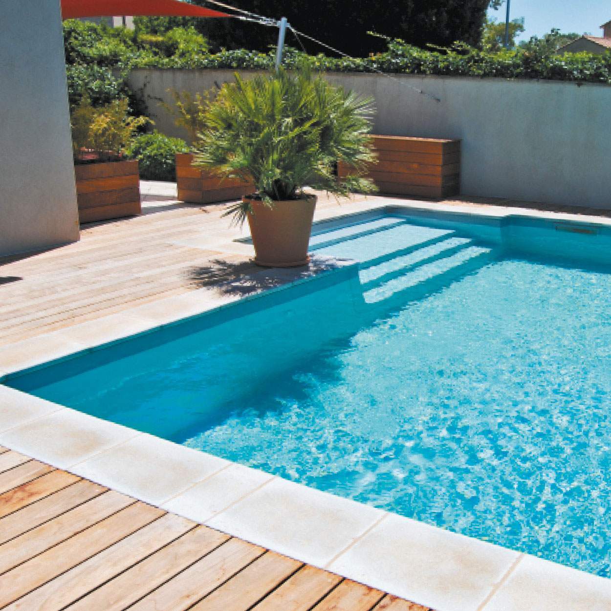 Piscine coque aux bords arrondis dans un espace extérieur aménagé avec terrasse en bois, eau turquoise, carrelage élégant, végétation verdoyante et éclairage naturel, créant une ambiance résidentielle relaxante.