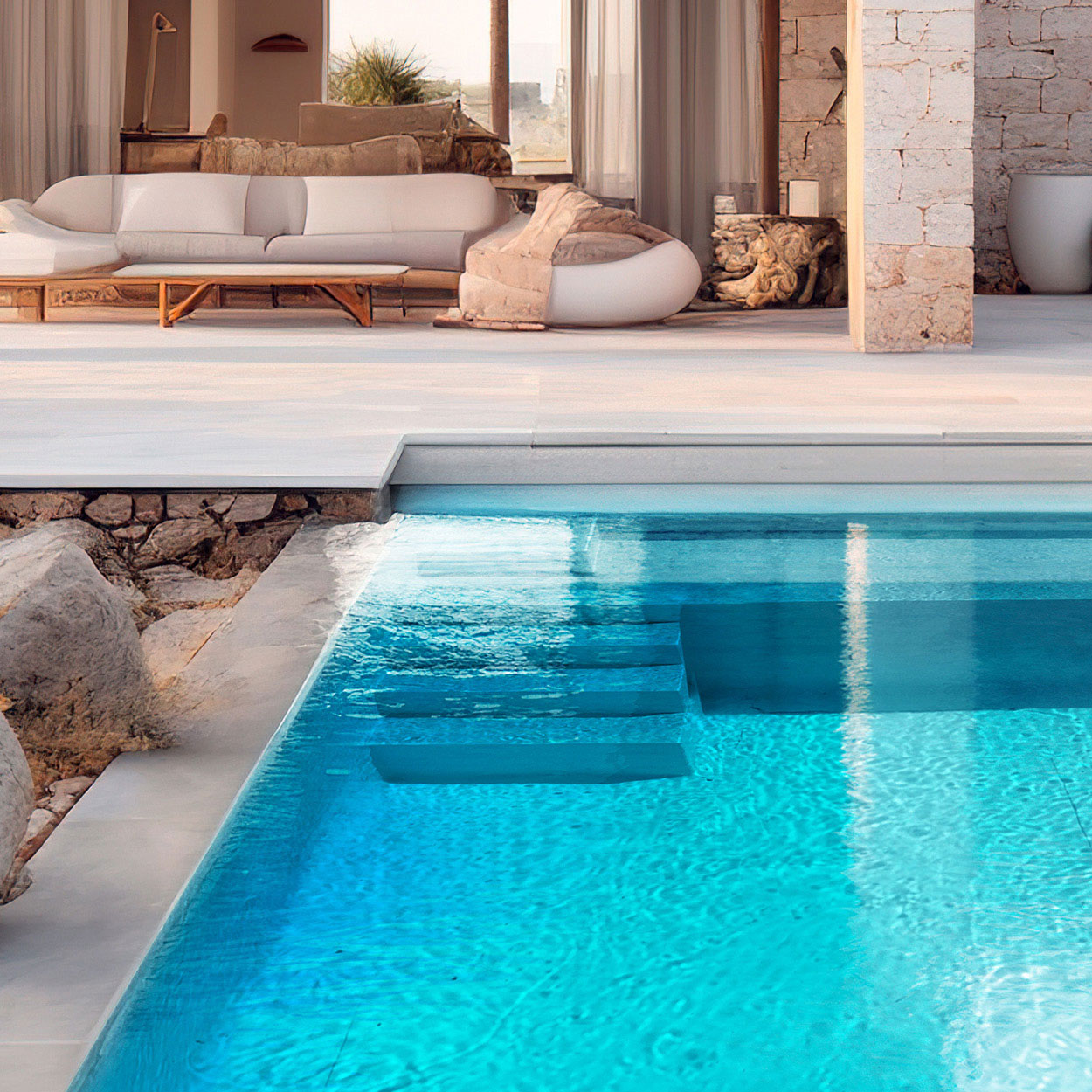 Piscine coque rectangulaire avec escaliers intégrés, bordée de dalles en pierre, entourée d'un espace de vie extérieur moderne avec canapé et poufs, lumière naturelle, éléments décoratifs en bois, intégration paysagère avec roches et végétation.