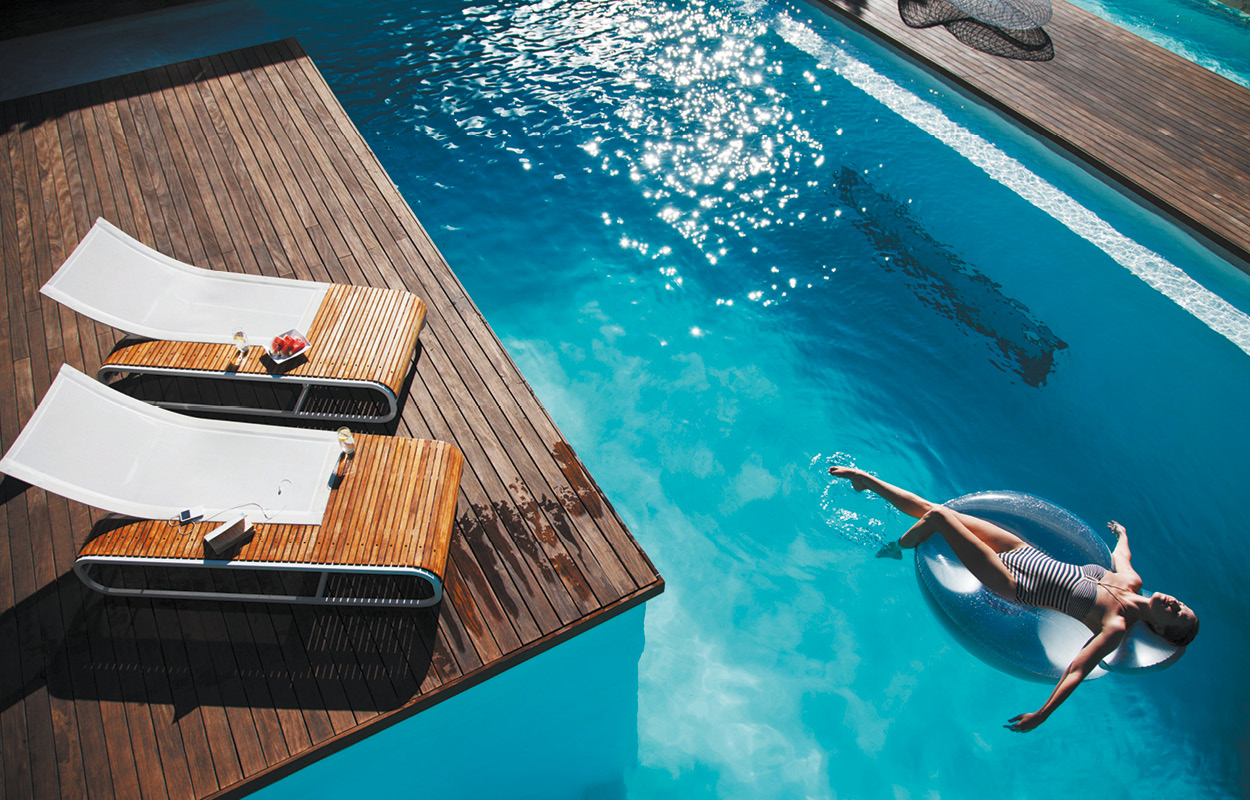 Scène estivale autour d'une piscine coque turquoise avec transats modernes, bouée transparente, et terrasse en bois, créant une ambiance de détente ensoleillée.